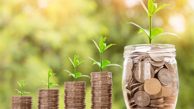 4 piles of incrementally growing coins from left to right, each with a correlating sprouting plant on top. Last in view is a glass jar containing the biggest amount of coins and the largest sprouting plant.